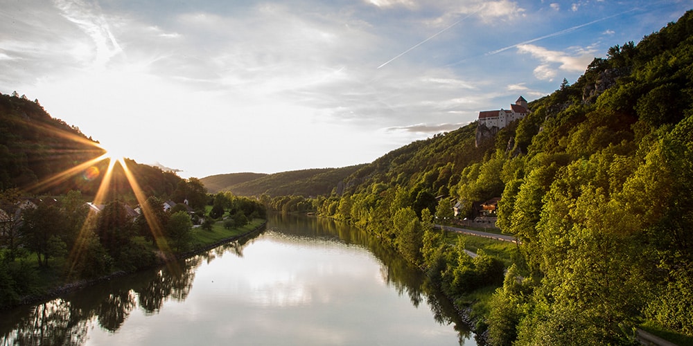 Foto mit Blick ins Altmühltal im Sonnenuntergang eingerahmt von Bäumen und der Burg Prunn